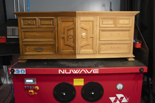 Wooden dresser on a Nuwave laser cleaning machine, with the left half showing the original darker finish and brass hardware, and the right half showing raw wood after laser cleaning with hardware removed